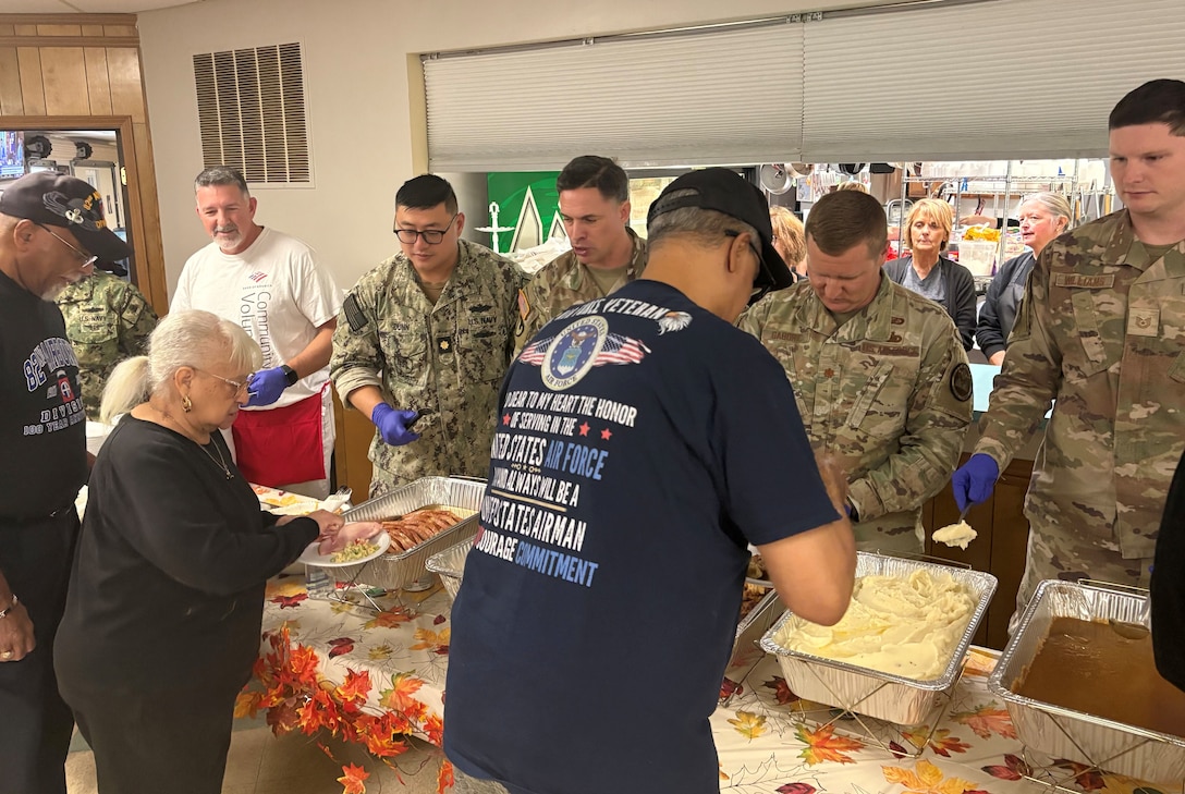 Several men in military uniform stand behind a table serving food to three people in the line.