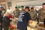 Several men in military uniform stand behind a table serving food to three people in the line.