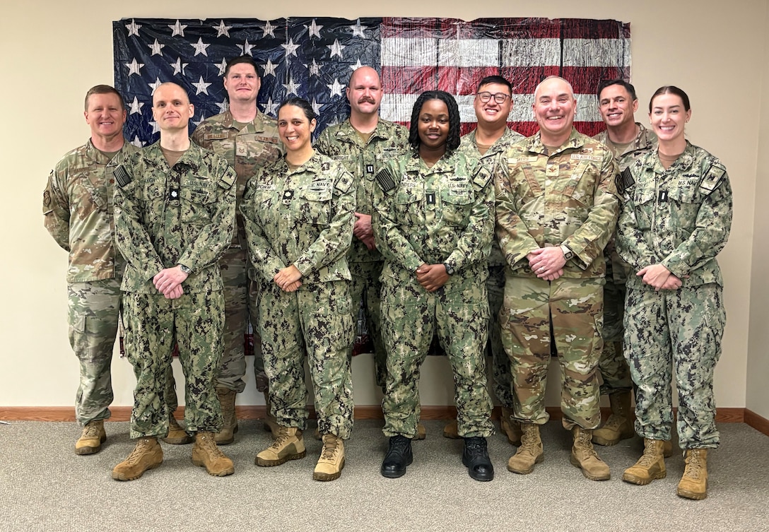 Ten people in military uniform stand posing for photo in front of a large American flag poster