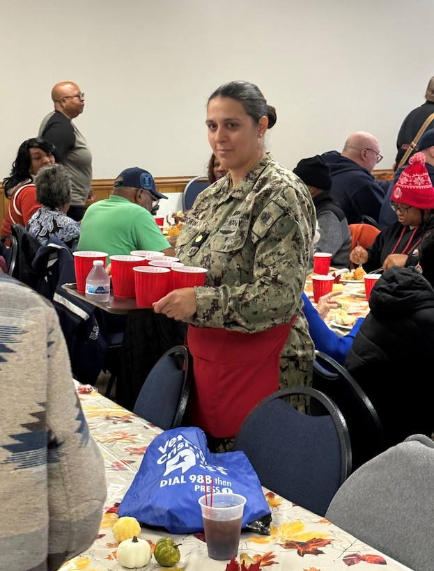 Woman in military uniform and red apron stands among tables with people seated, holding a tray with red cups.