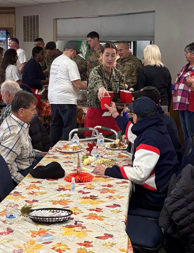 Woman in military uniform and red apron serves drinks in red cups to people seated at table covered in fall leaves designed table cloth.