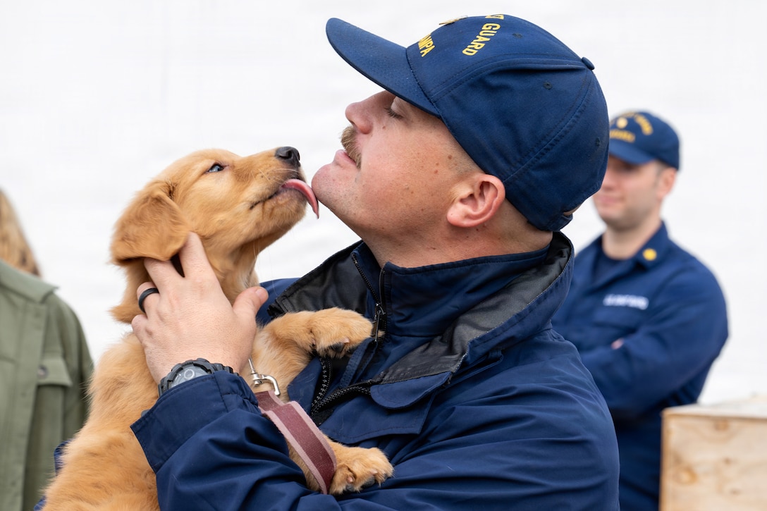 A Coast Guardsman standing outdoors holds a puppy that licks his chin.