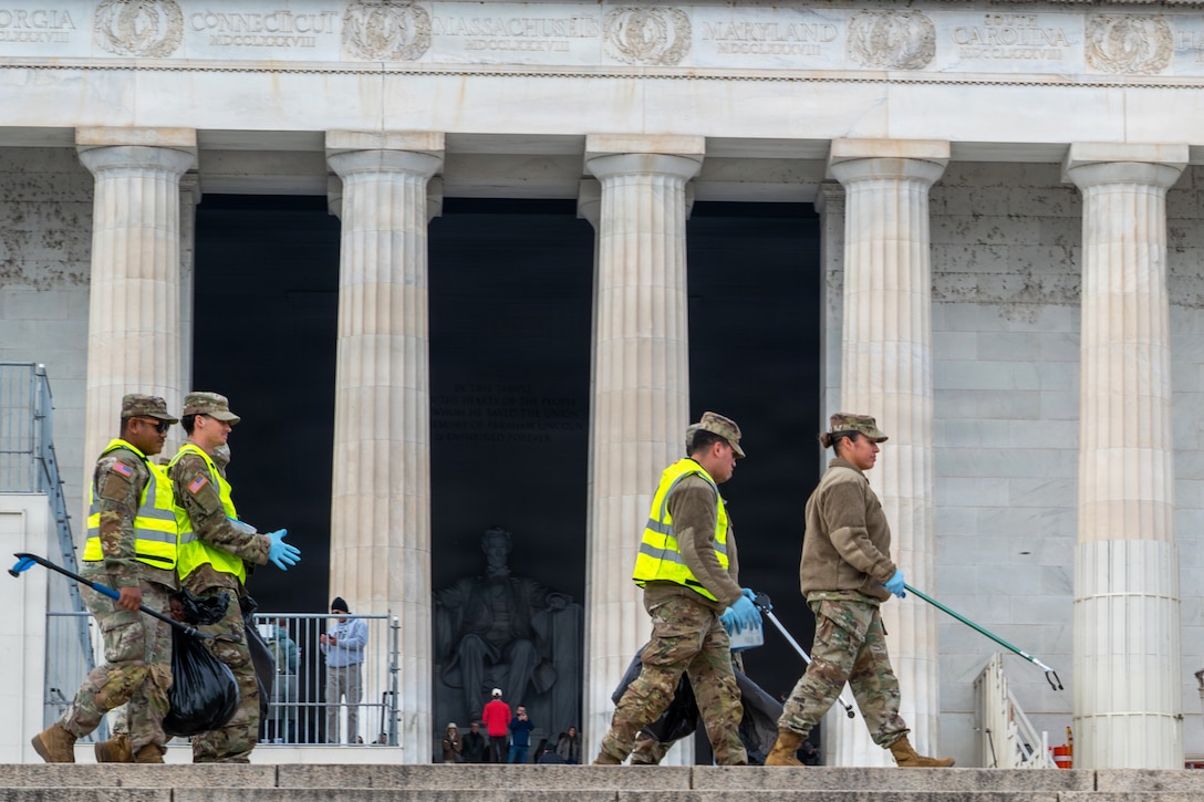 Guardsmen carrying trash bags and devices to pick up trash walk in front of a large monument with five columns and a statue of a person sitting in a chair, with other people standing in the background closer to the monument.