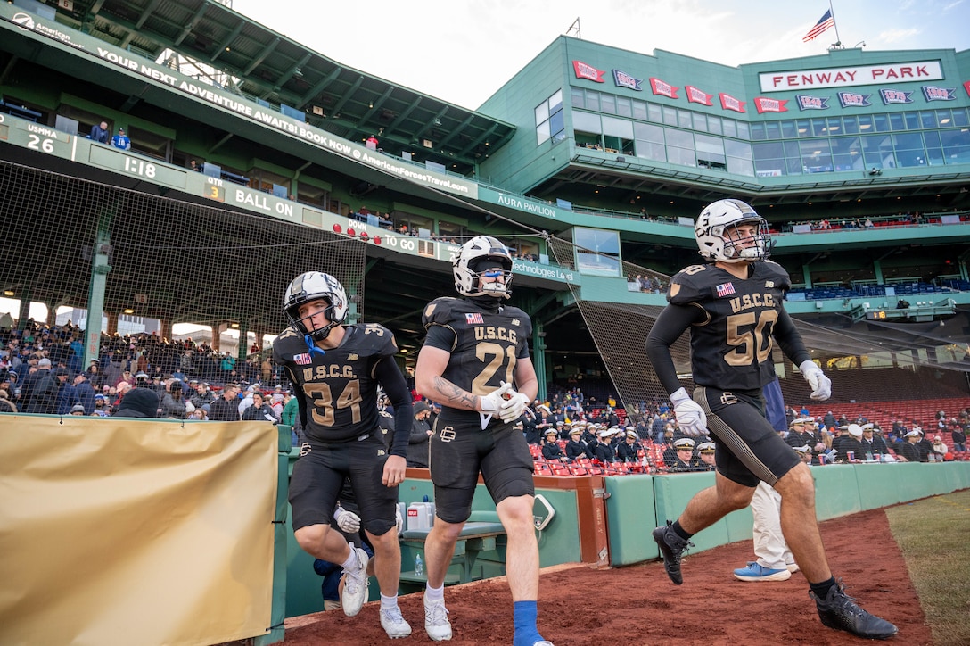 U.S. Coast Guard Academy (USCGA) Cadet Fourth Class Cooper Price (left to right), Cadet Third Class Cal Schubert, and Cadet Fourth Class JT Nelly run onto the field following the conclusion of the halftime show at Fenway Park, Boston, Nov. 15, 2025. USCGA won last year's Secretaries' Cup, ending the game with a 42-21 score against the U.S. Merchant Marine Academy. (U.S. Coast Guard photo by Petty Officer Third Class Leo Avila.)