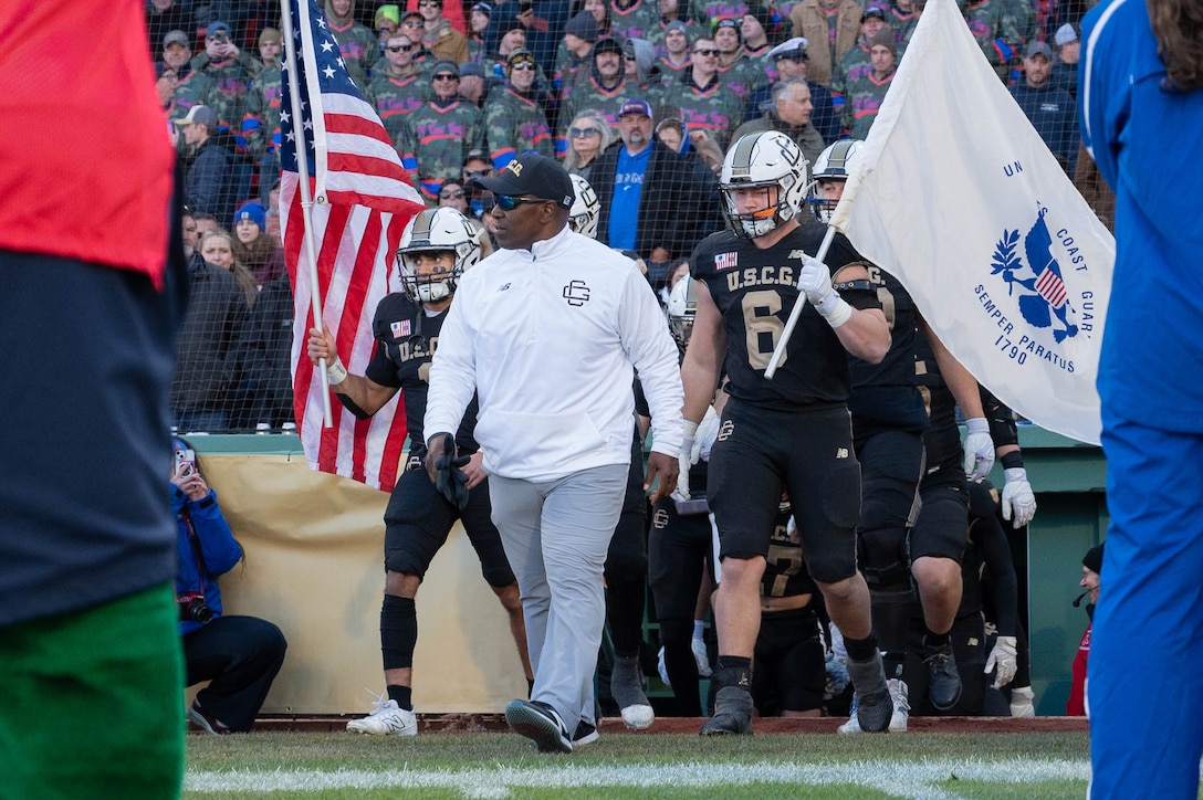 U.S. Coast Guard Academy (USCGA) football head coach C.C. Grant (middle, white sweater) walks onto the field with his football team during the 54th Secretaries' Cup at Fenway Park, Boston, Nov. 15, 2025. Grant has been a coach at USCGA since 1998 and became head coach in 2020. (U.S. Coast Guard photo by Petty Officer Third Class Leo Avila.)
