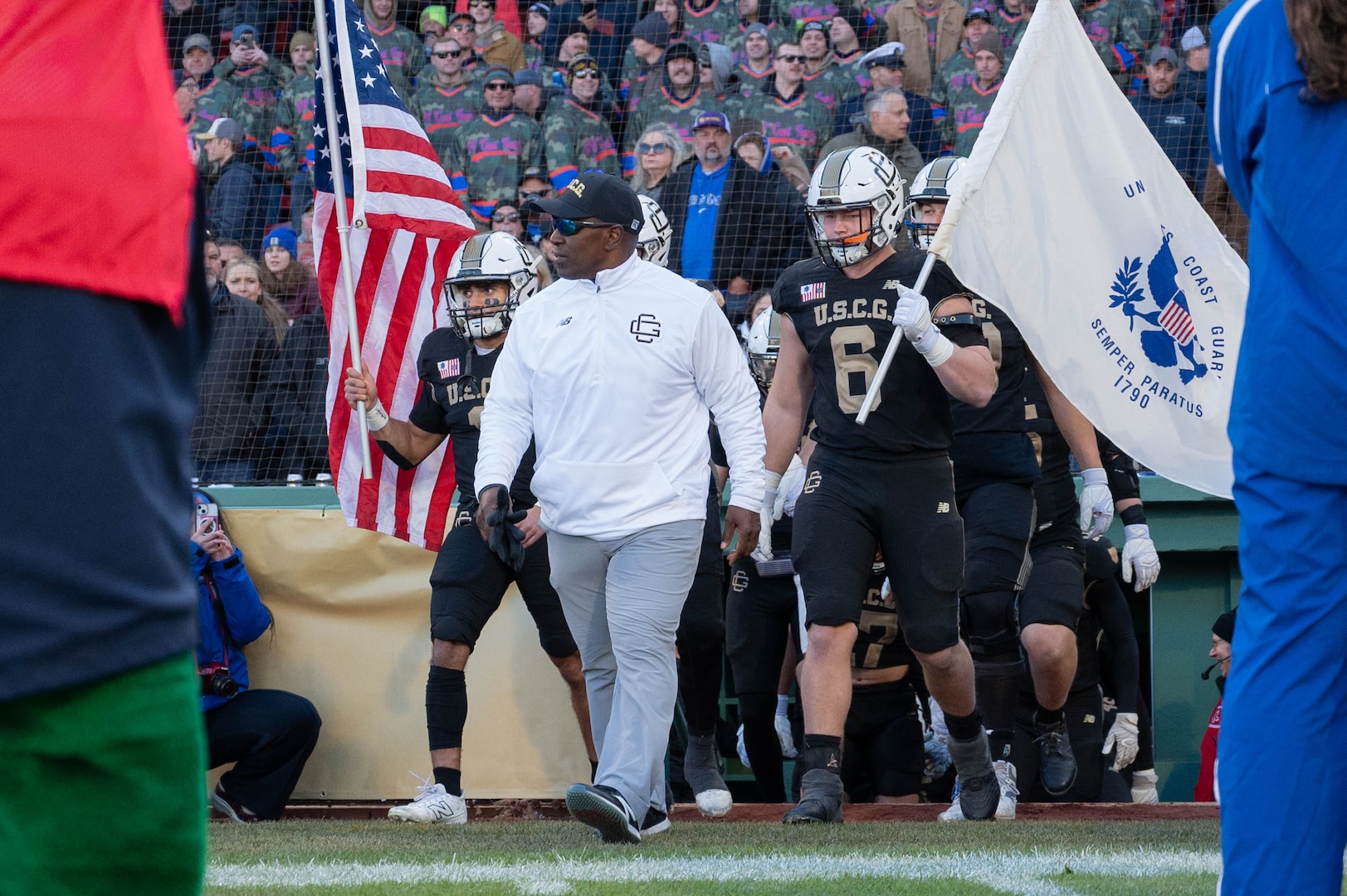 U.S. Coast Guard Academy (USCGA) football head coach C.C. Grant (middle, white sweater) walks onto the field with his football team during the 54th Secretaries' Cup at Fenway Park, Boston, Nov. 15, 2025. Grant has been a coach at USCGA since 1998 and became head coach in 2020. (U.S. Coast Guard photo by Petty Officer Third Class Leo Avila.)