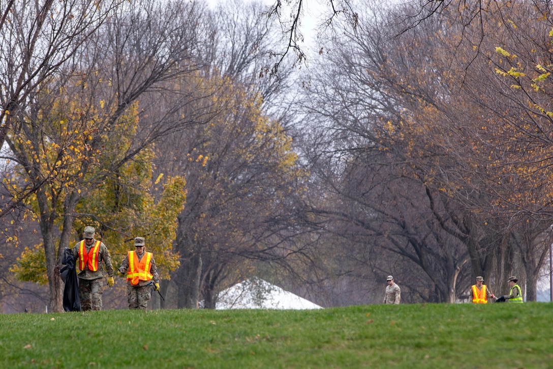 Five guardsmen wearing bright reflective vests collect trash in a field surrounded by leafless trees on a gloomy day.