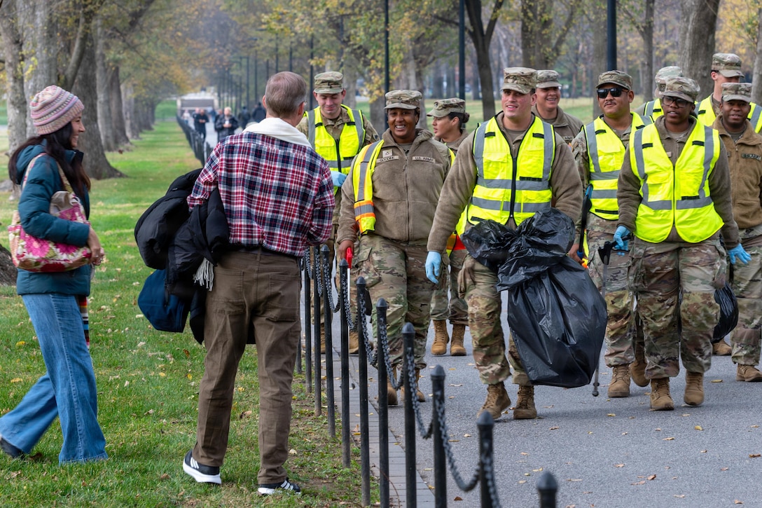 Nearly a dozen guardsmen wearing reflective vests walk in formation on the pavement while interacting with two tourists standing on grass.