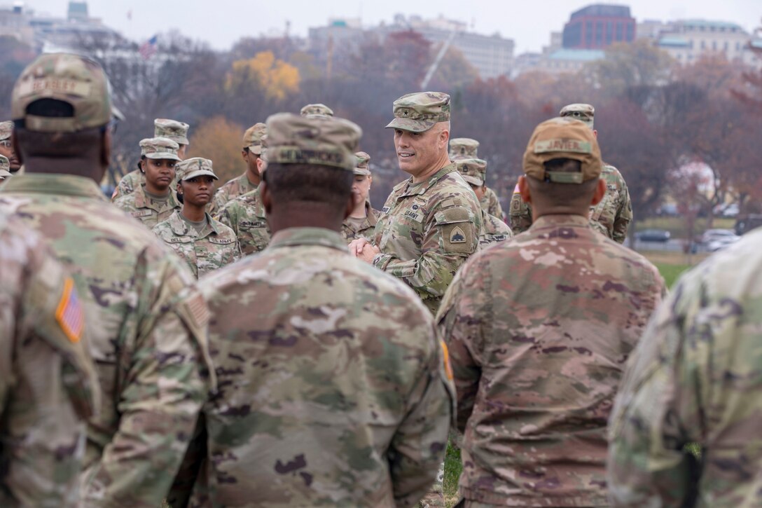 A commander speaks in front of dozens of guardsmen with a city view in the background on a gloomy day.