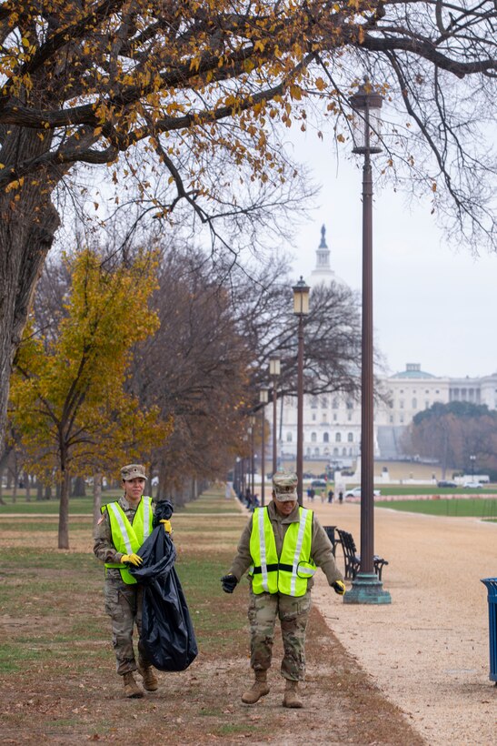Two guardsmen both wearing reflective vests and one carrying a trash bag walk in front of a building on a gloomy day.