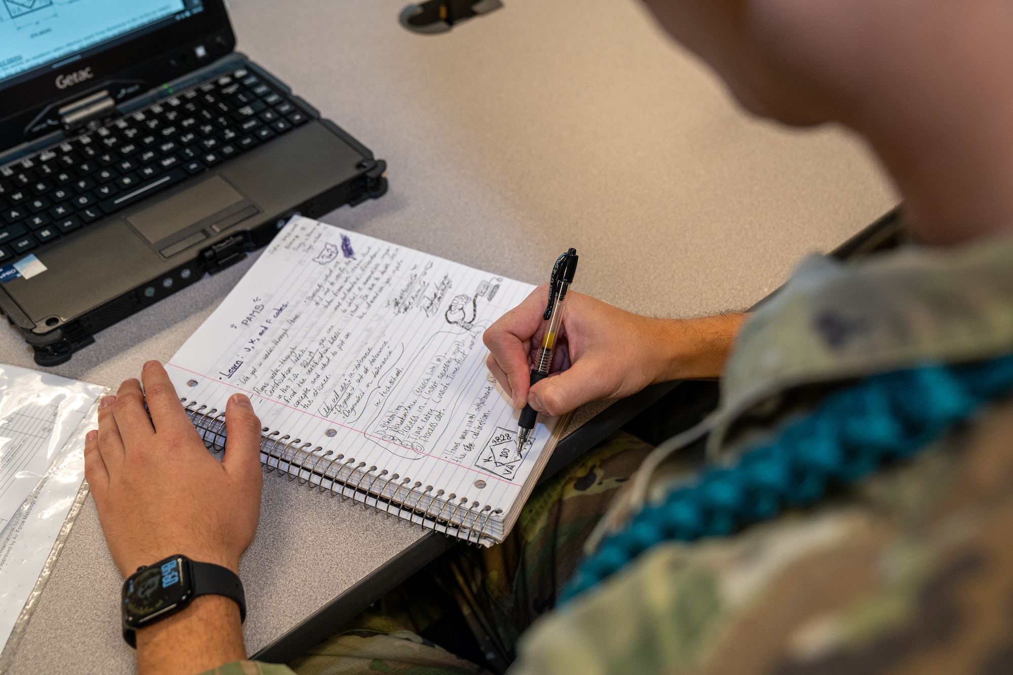A serviceman in OCPs wearing a teal aiguillette writes down notes in a notebook.