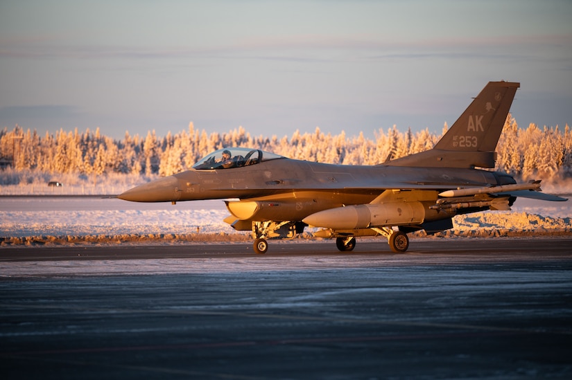 A military fighter jet sits on an airport taxiway. Snow is covering the ground and trees in the background.