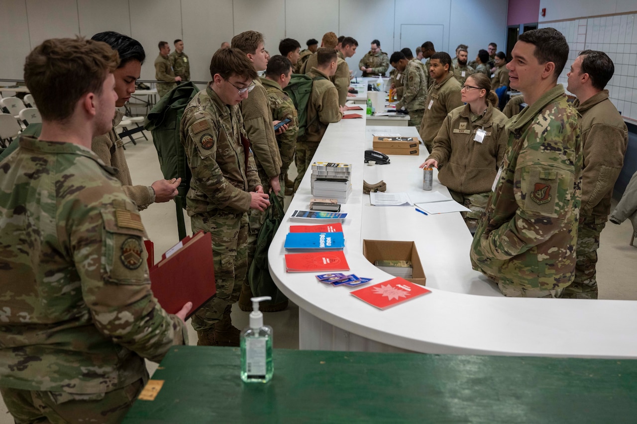 Dozens of people wearing camouflage military uniforms stand around a counter talking. There are several different books lying on the counter and some of the service members are holding folders.