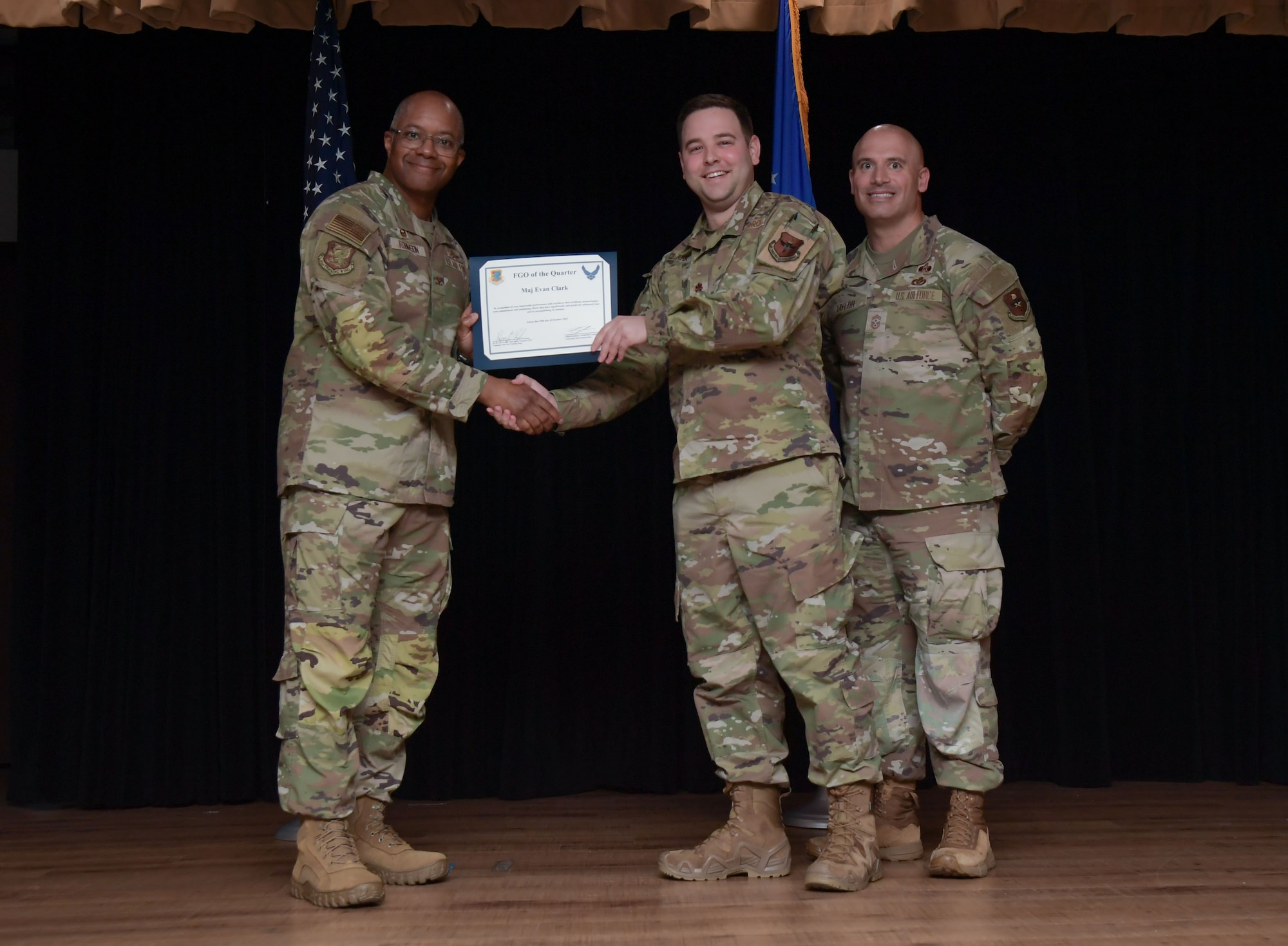Three men wearing military uniforms stand on stage with the center man holding a certificate