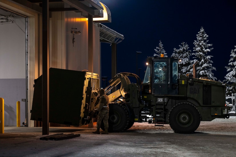 A man driving a forklift moves a large container into a building while another man walks alongside the forklift outside. There are trees in the background with snow on them.
