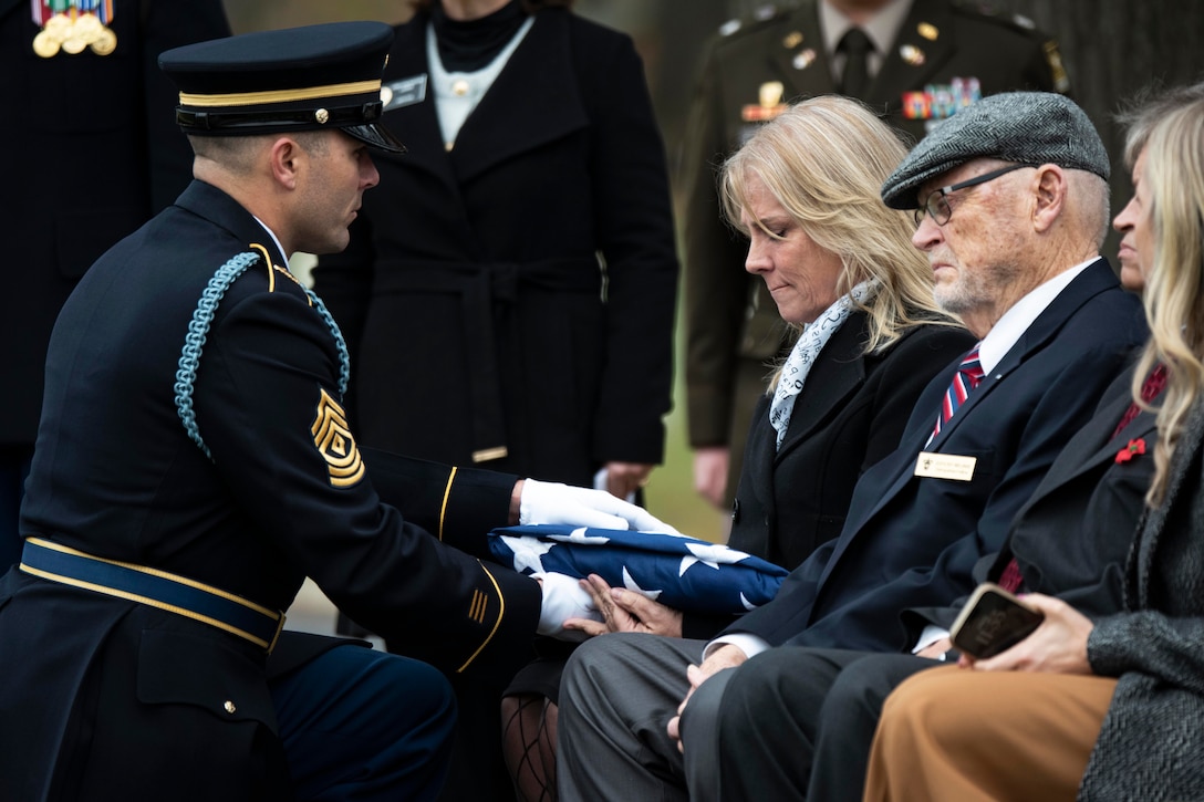 A service member in a blue uniform kneels to hand a folded American flag to a person sitting in a chair  outdoors, with other people sitting nearby and others standing in the background.