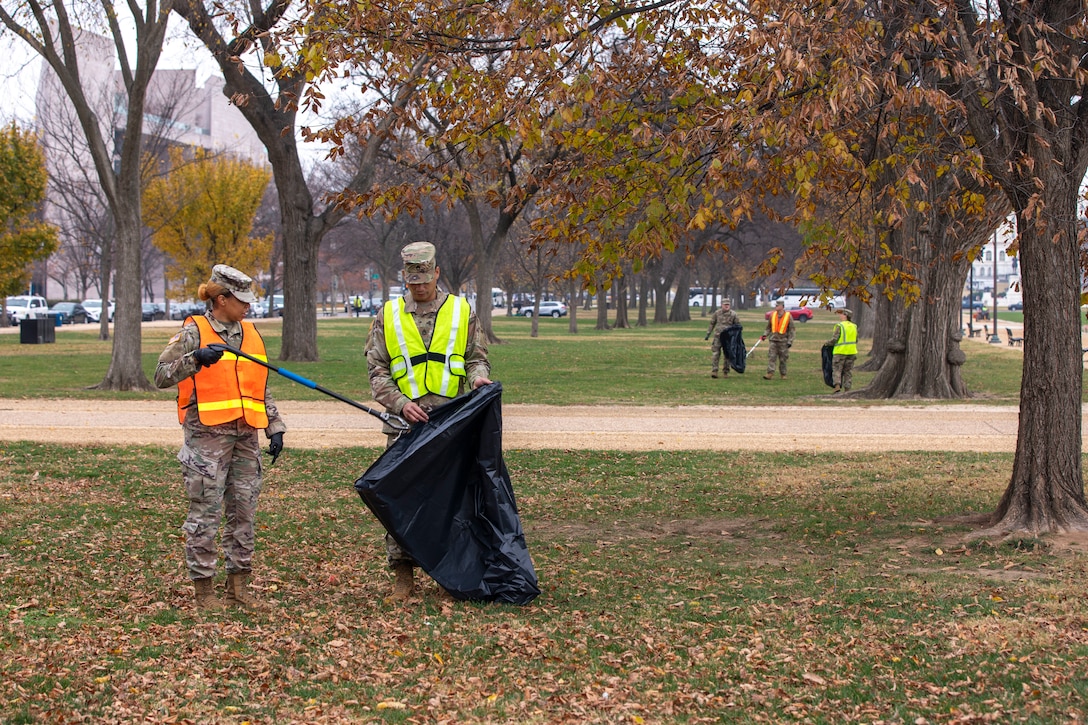 A guardsman puts trash into a bag as fellow guardsmen collect trash in a field in the background on a gloomy day.