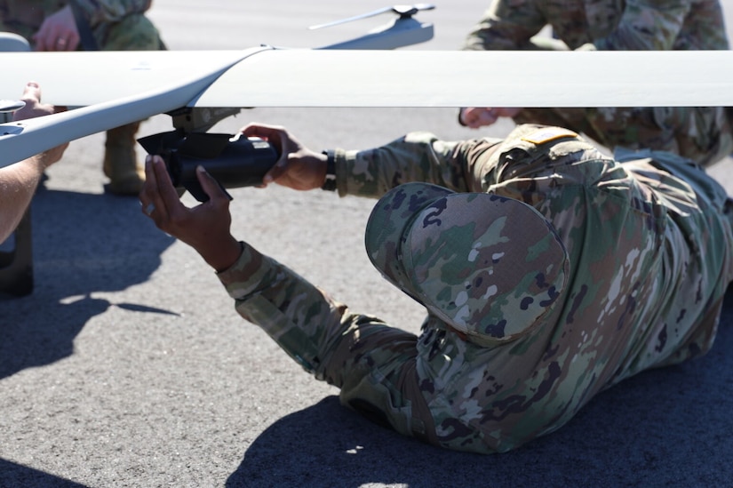A person in a camouflage military uniform lies on the ground outside, under the wing of a small aircraft, to attach an item.