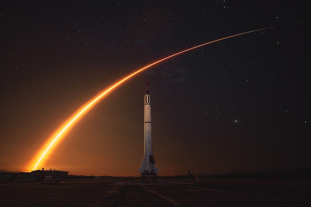 A rocket leaves a stream of orange light in a dark sky after launch, with an upright rocket seen on a runway in the foreground with "United States" written on it.