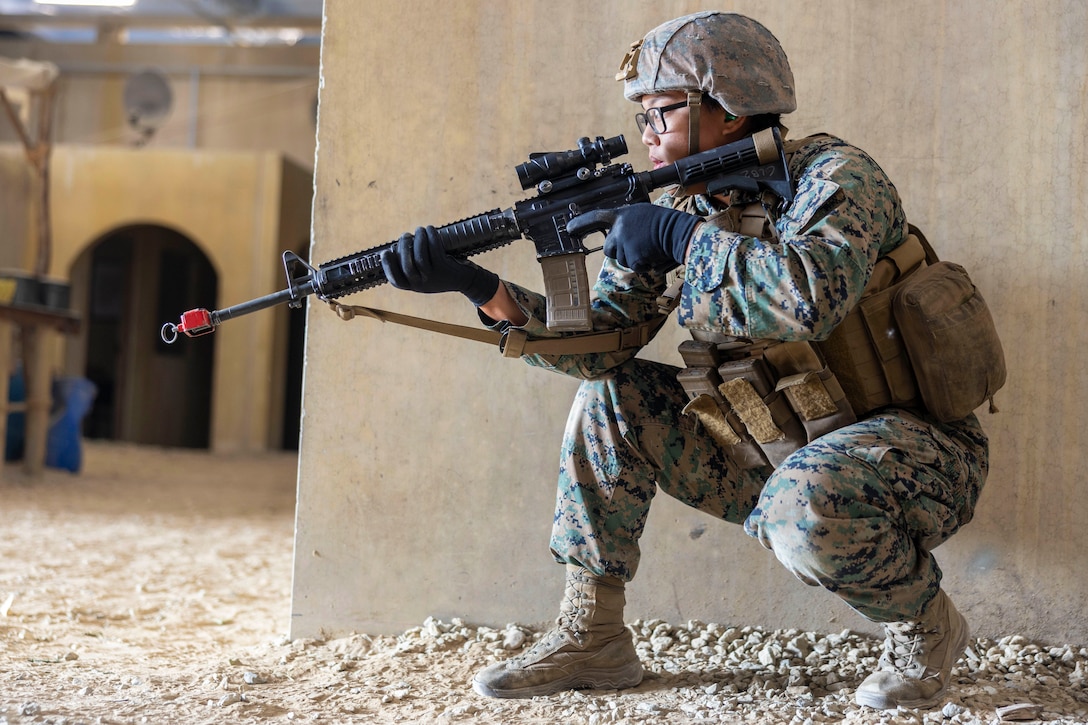 A Marine in combat gear and holding a machine gun crouches near a wall in a building-like setting on rocky ground.