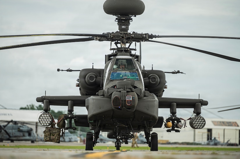 A pilot sits in the cockpit of a military helicopter on the tarmac at a military installation. The aircraft is carrying counter-unmanned aerial system weapons.