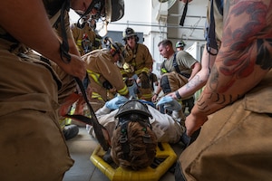An Airman lies on a litter while firefighters kneel around him strapping him to the litter
