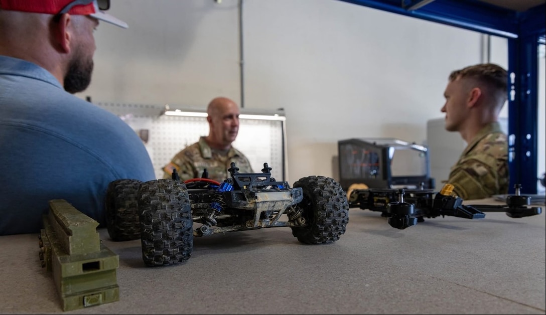 Three men meet with various military models and tools on a table.