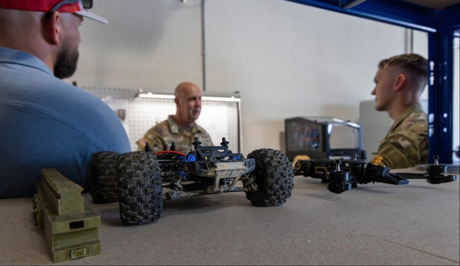 Three men meet with various military models and tools on a table.