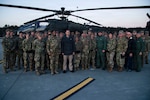 A group of service members in uniform and civilians stand in front of a military helicopter.