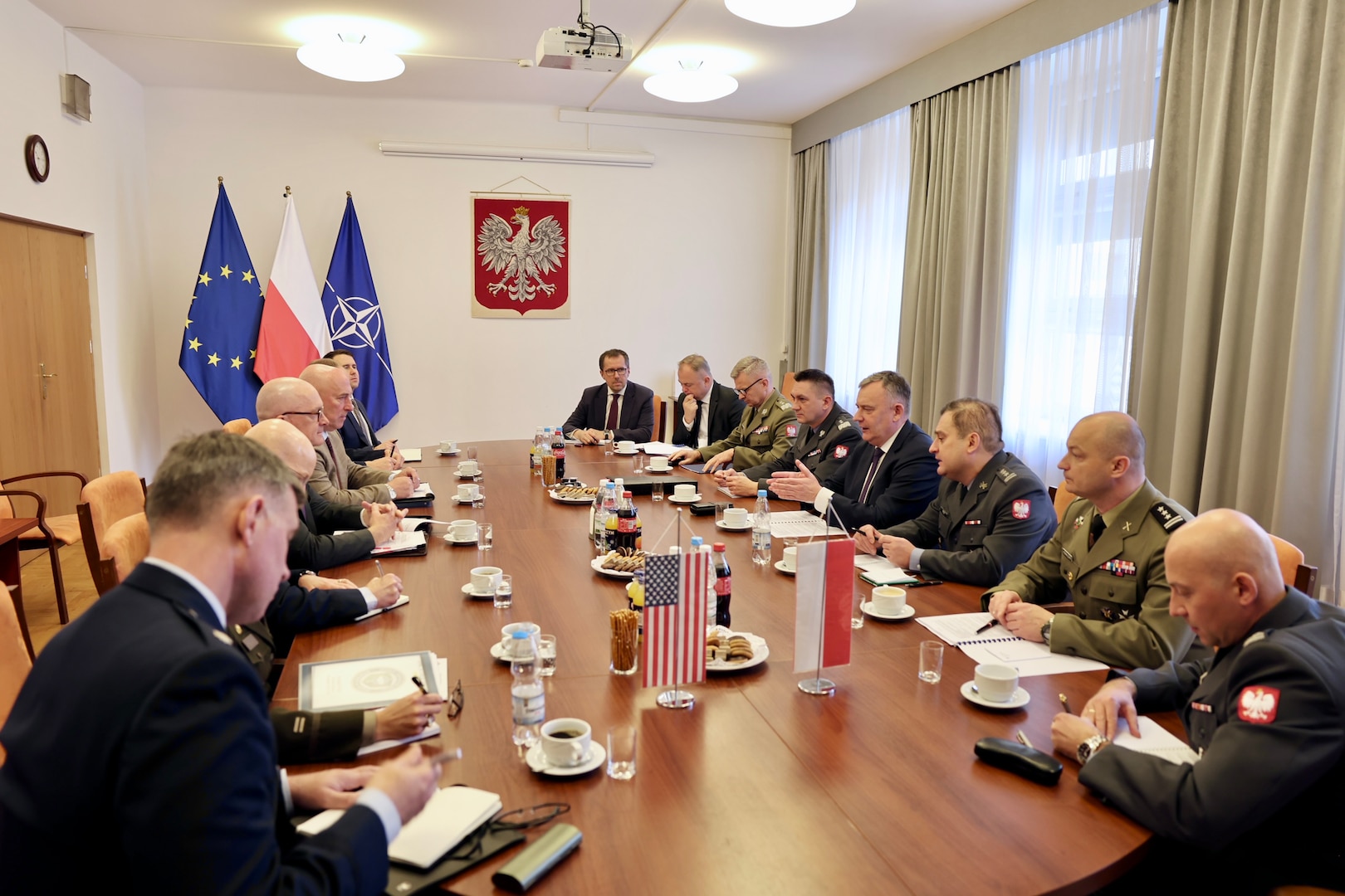 A group of people sit at conference table with flags in the background.