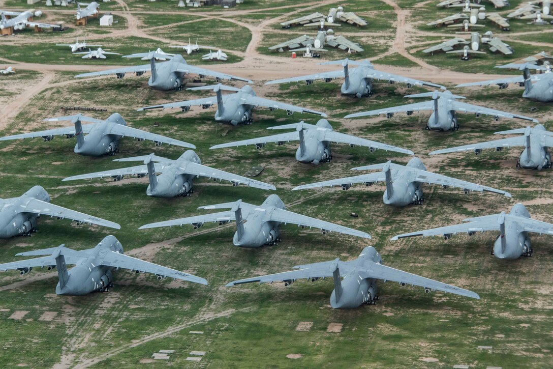 Lines of planes are parked on grass during the day as seen from above.