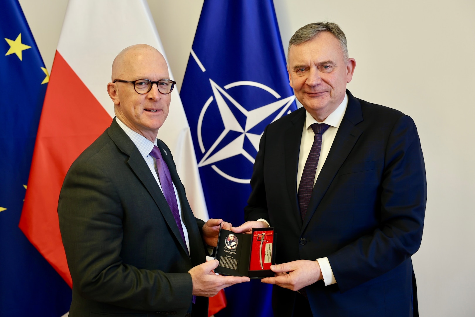 Two men hold a small gift box while posing for a photo in front of flags.