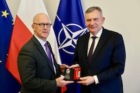 Two men hold a small gift box while posing for a photo in front of flags.