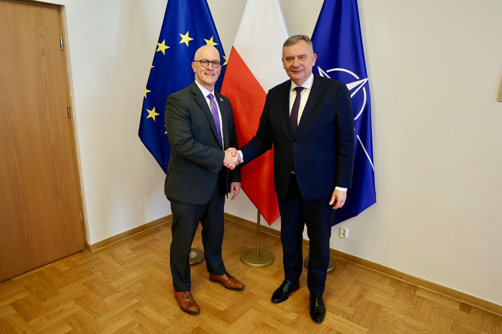 Two men shake hands while posing for a photo in front of flags.