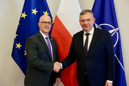 Two men shake hands while posing for a photo in front of flags.