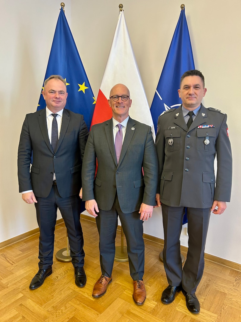 Three people stand in front of flags while posing for a photo.