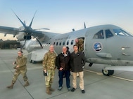 Four people stand on a tarmac in front of a military aircraft.