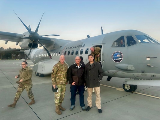 Four people stand on a tarmac in front of a military aircraft.