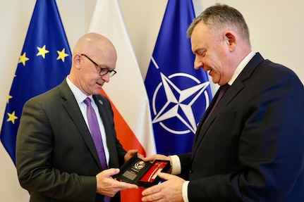 Two people in business suits hold a small gift box while standing in front of flags.