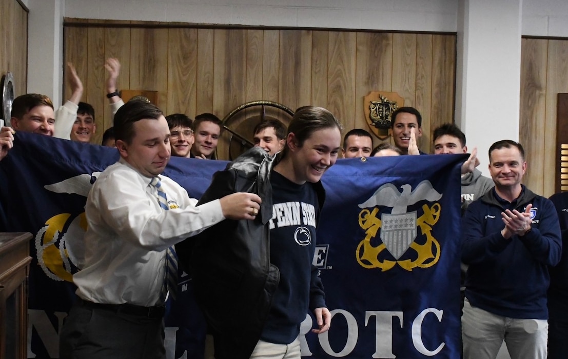 A Midshipman, a senior at Penn State University, dons her Surface Warfare Officer (SWO) jacket after selecting her first ship assignment during the bi-annual ship selection event coordinated by Navy Personnel Command PERS-41 SWO Assignments. Naval Reserve Officers' Training Corps (NROTC) is under the charge of Naval Service Training Command (NSTC), which oversees 98 percent of the Navy’s officer and enlisted accessions. NSTC, headquartered at Naval Station Great Lakes, Illinois, is the Foundry of America’s Fighting Force. NSTC transforms America’s sons and daughters into service professionals through Recruit Training Command, Officer Training Command Newport, and NROTC. Additionally, NSTC leads the Navy Junior ROTC citizenship development program, which instills leadership, character, and civic responsibility in high school students globally.