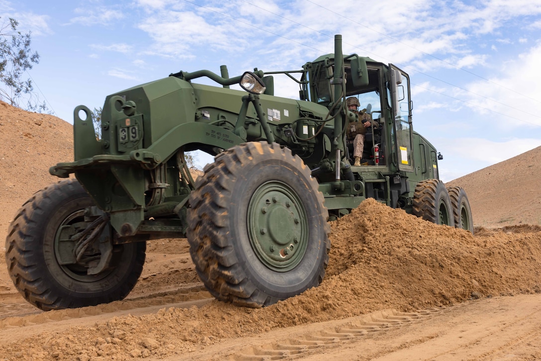 A Marine operates a green motorized road grader in the dirt under a cloudy, blue sky.