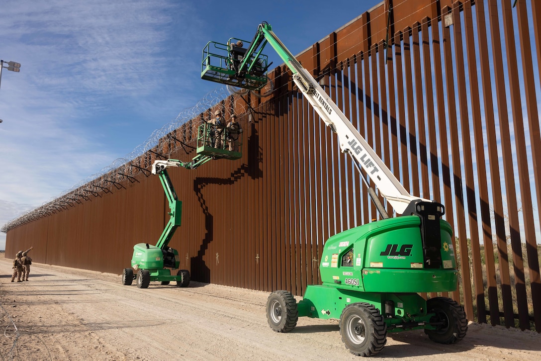 Marines on two green lifts attach concertina wire to a large metal fence in a desert-like area.