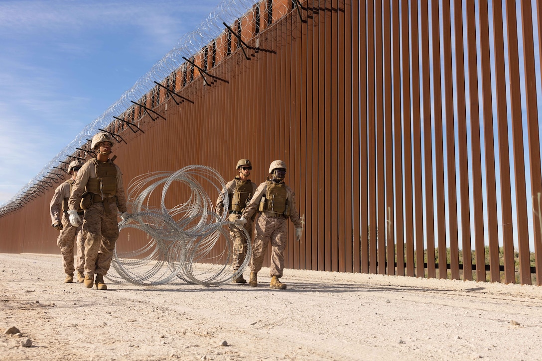 Four Marines in protective gear carry concertina wire while walking next to a large metal fence in a desert-like area.
