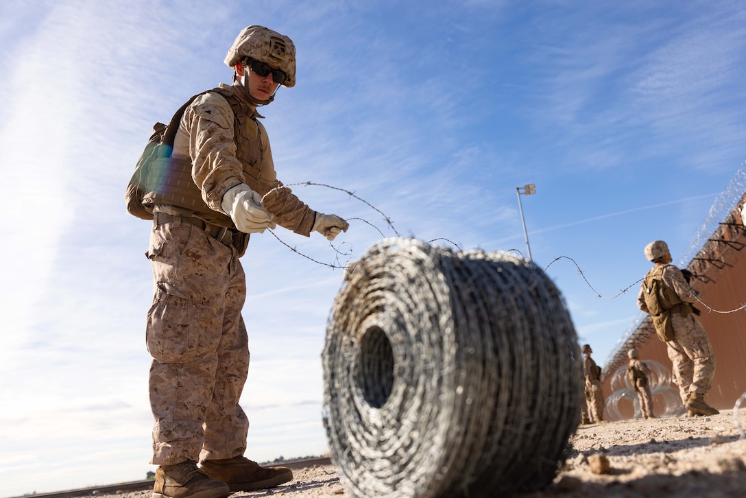 A Marine unravels a wheel of wire as fellow Marines attach it to the top of a metal fence in the background in a desert-like area.