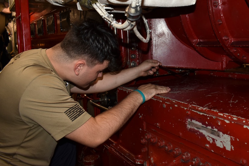 A man wearing a tan T-shirt works in the engine room of a military submarine.
