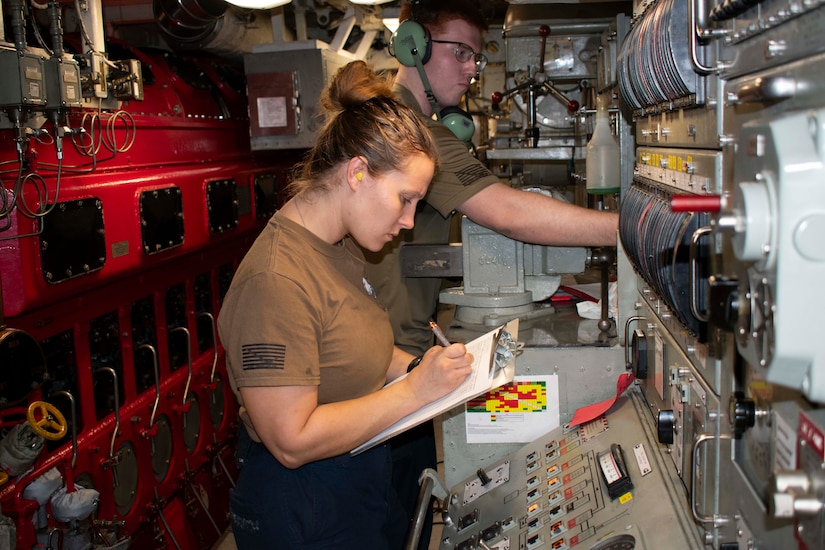 A woman wearing a tan T-shirt writes on a piece of paper attached to a clipboard while standing in the engine room of a military submarine. A man in similar attire works behind her.