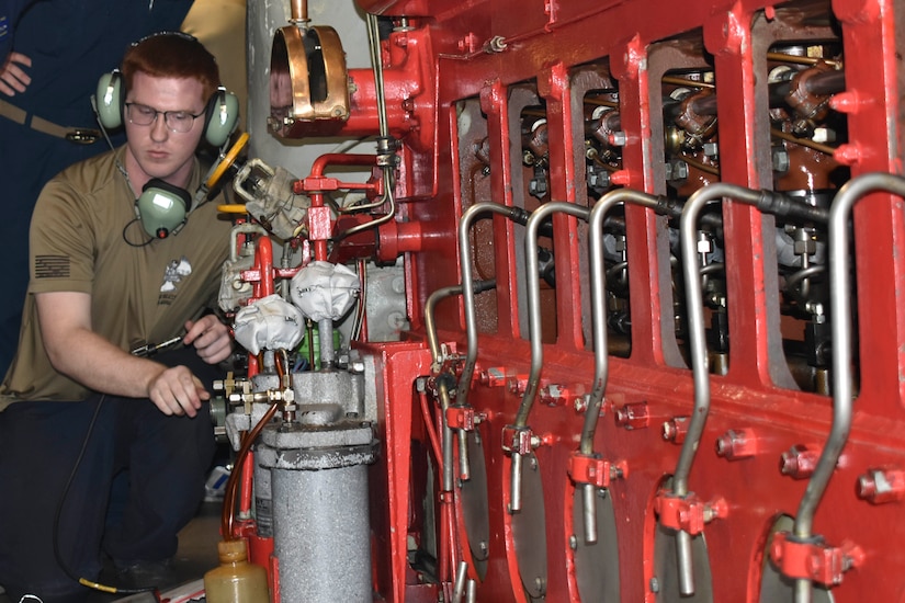 A man wearing a tan T-shirt opens a valve in the engine room of a military submarine.