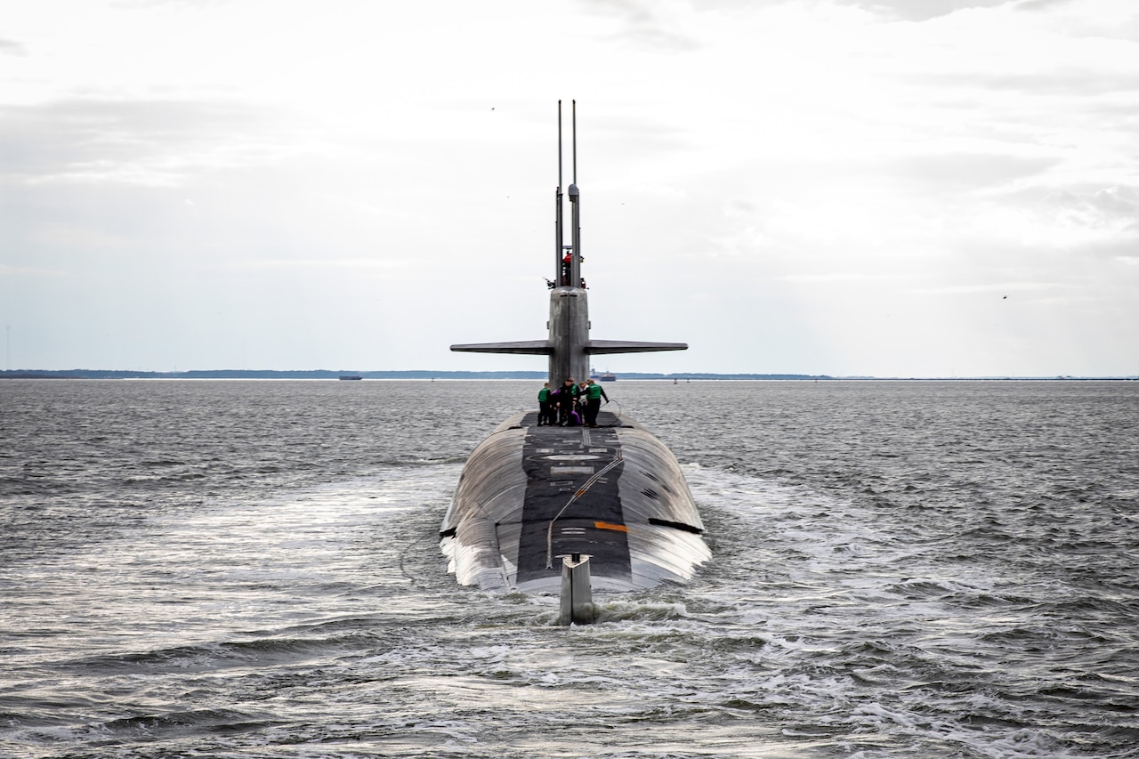 A military submarine sails on the surface of the water. There are several service members working on top of the vessel.