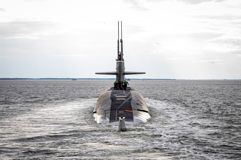 A military submarine sails on the surface of the water. There are several service members working on top of the vessel.