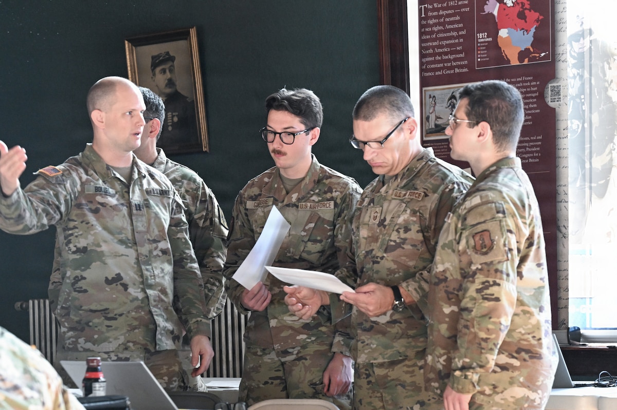 A group of uniformed service members stand together talking and reviewing paperwork by a bright window.